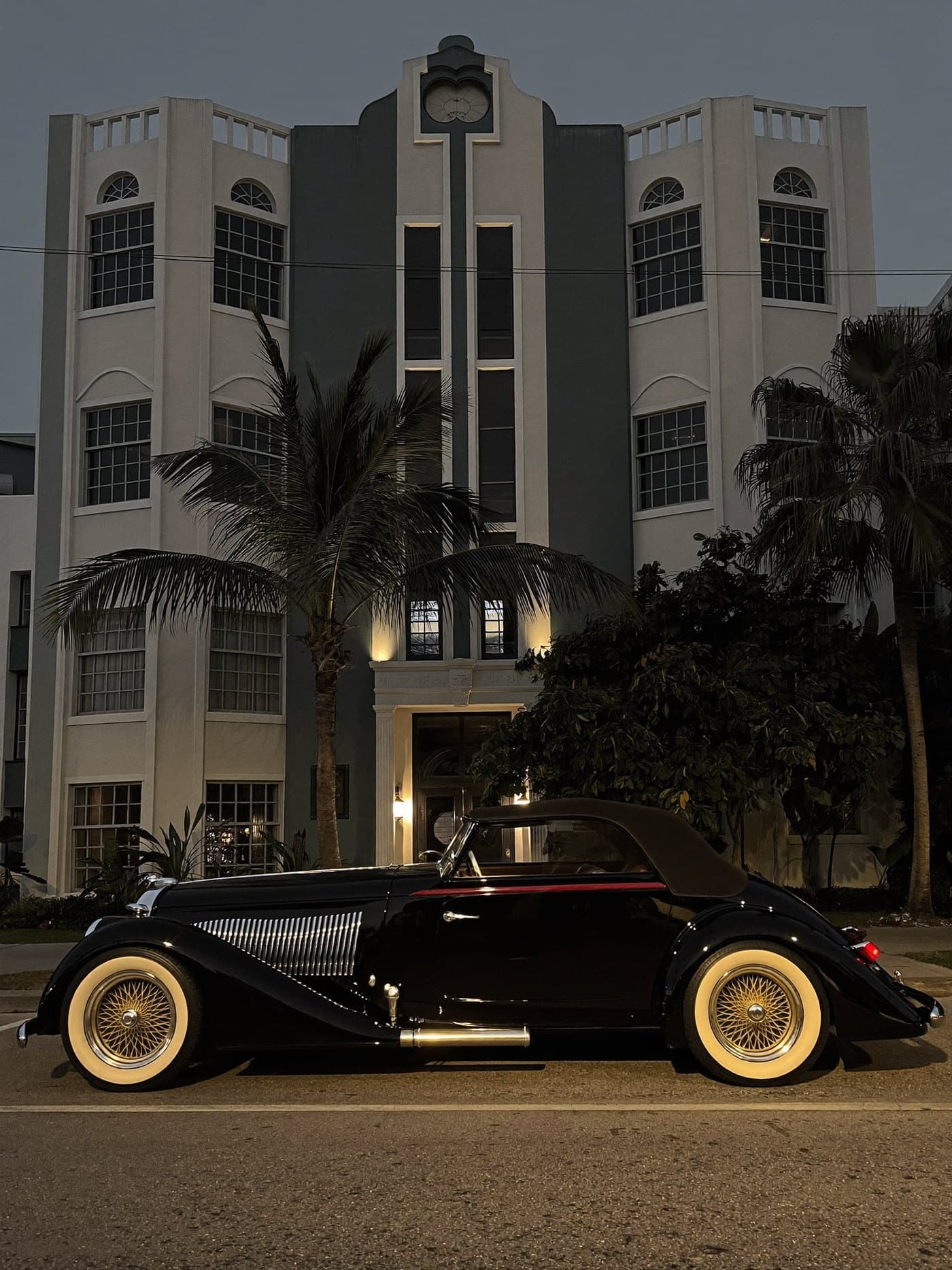 A vintage black grand touring car outside an art deco building at dusk