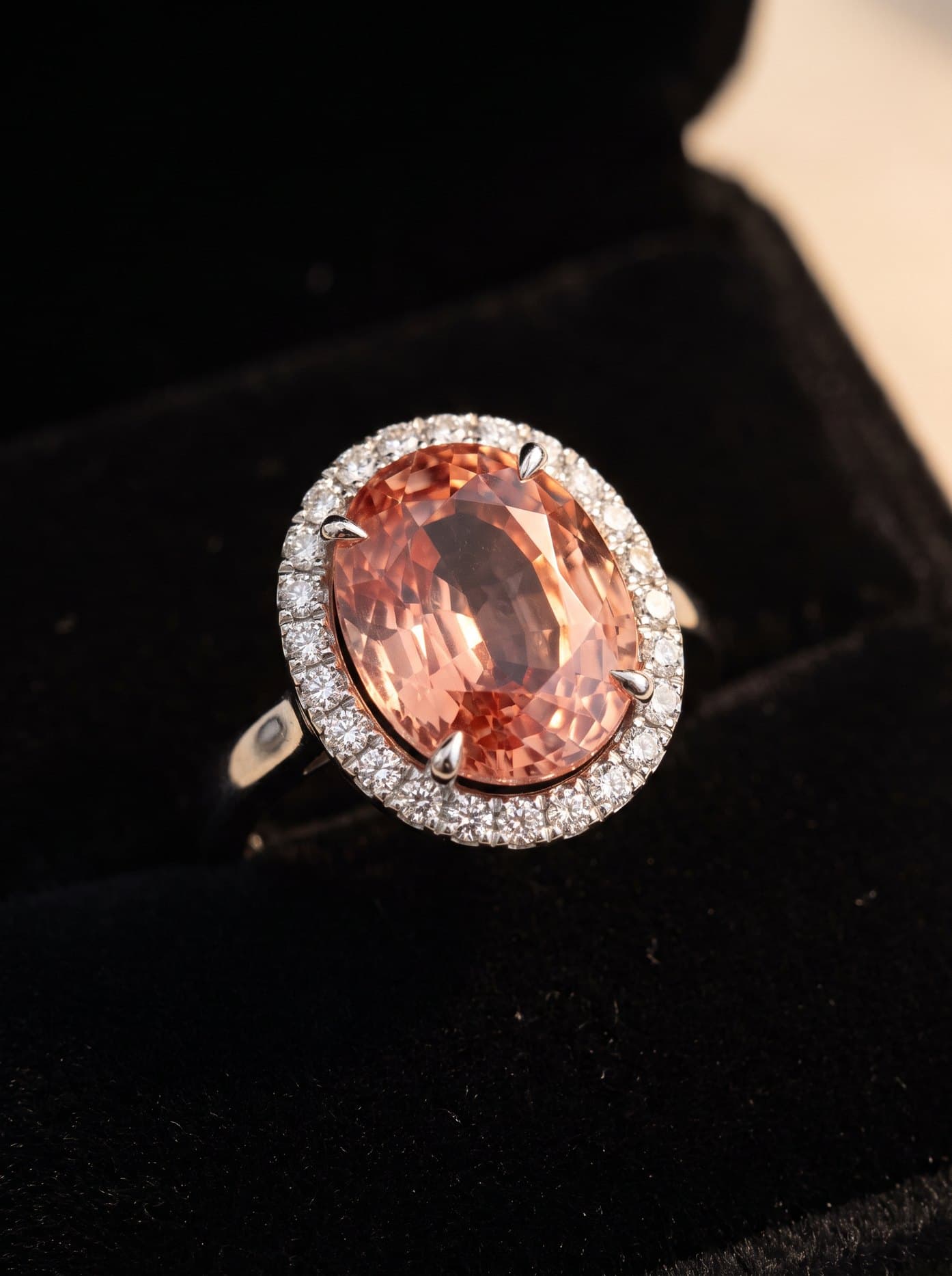 An oval padparadscha sapphire ring with a diamond halo on black velvet